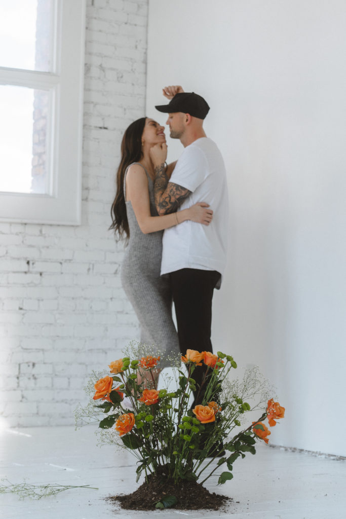 Young creative couple taking their engagement photos in an all white painted studio with creative floral prop 